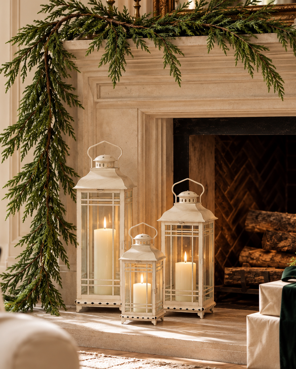 A festive holiday living room scene featuring a green evergreen garland draped over a cream-colored stone fireplace mantel, accompanied by three white lanterns with lit candles on the hearth. A beige sofa with a green pillow is visible on the left, and wrapped gift boxes sit on the right.